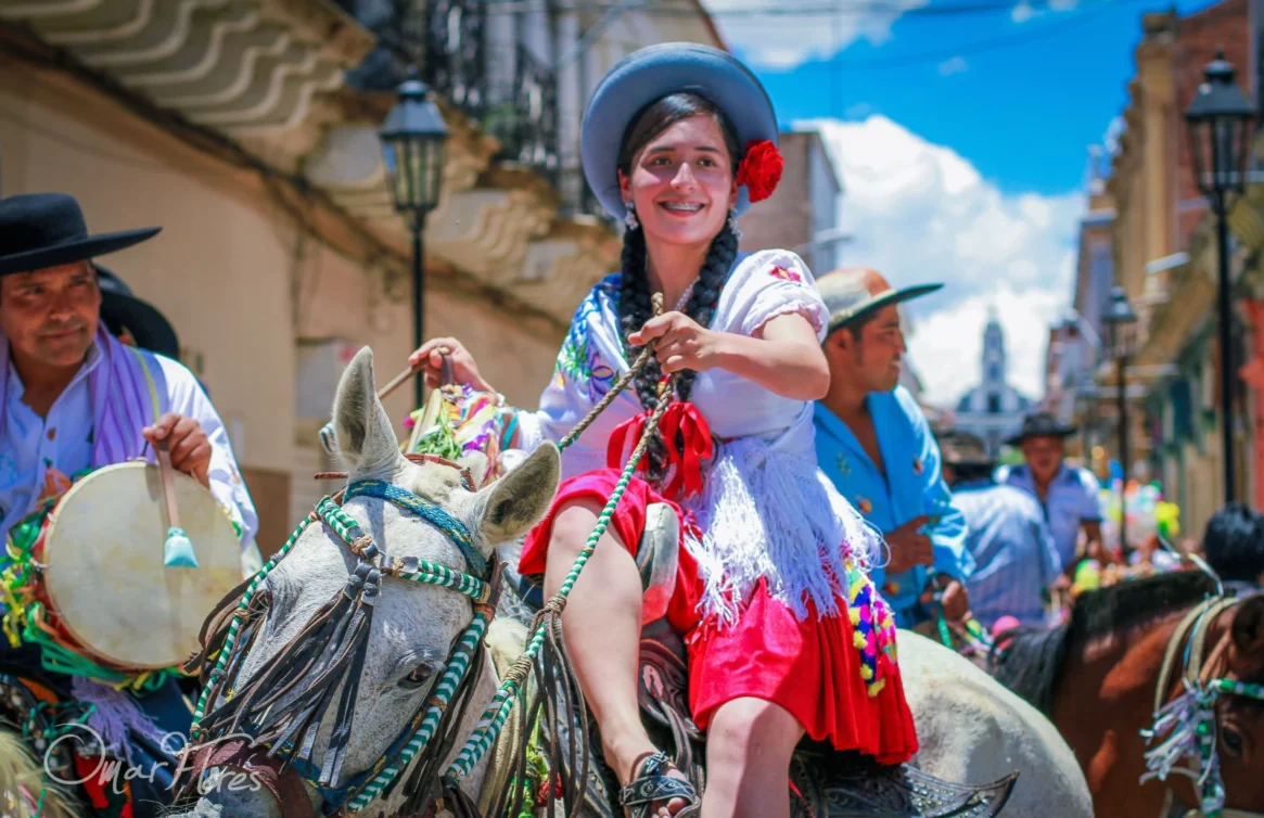 Desfile em Tarija com uma mulher em traje tradicional montada em um cavalo e músicos atrás dela.