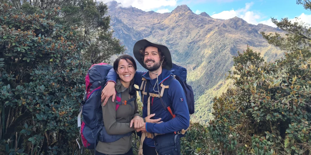 Pareja de senderistas sonrientes en el Camino Inca, rodeados de vegetación y montañas andinas al fondo.