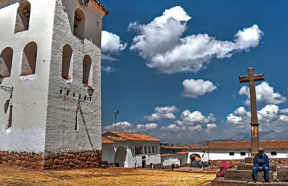 Torre branca e cruz na praça de Chinchero, com montanhas ao fundo.