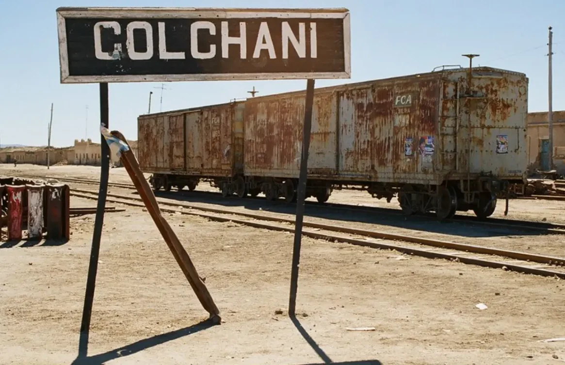 Placa de Colchani ao lado de vagões enferrujados na ferrovia do Altiplano boliviano.