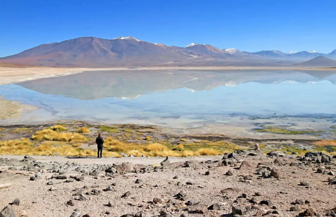 Pessoa em frente à Laguna Blanca com montanhas refletidas na água, na Bolívia.