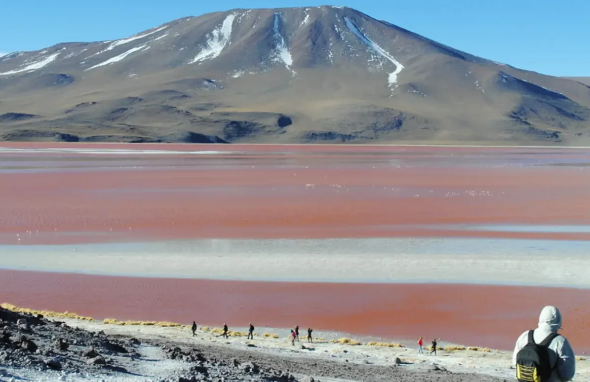 Persona observa laguna altiplánica rojiza con montaña al fondo en Bolivia.