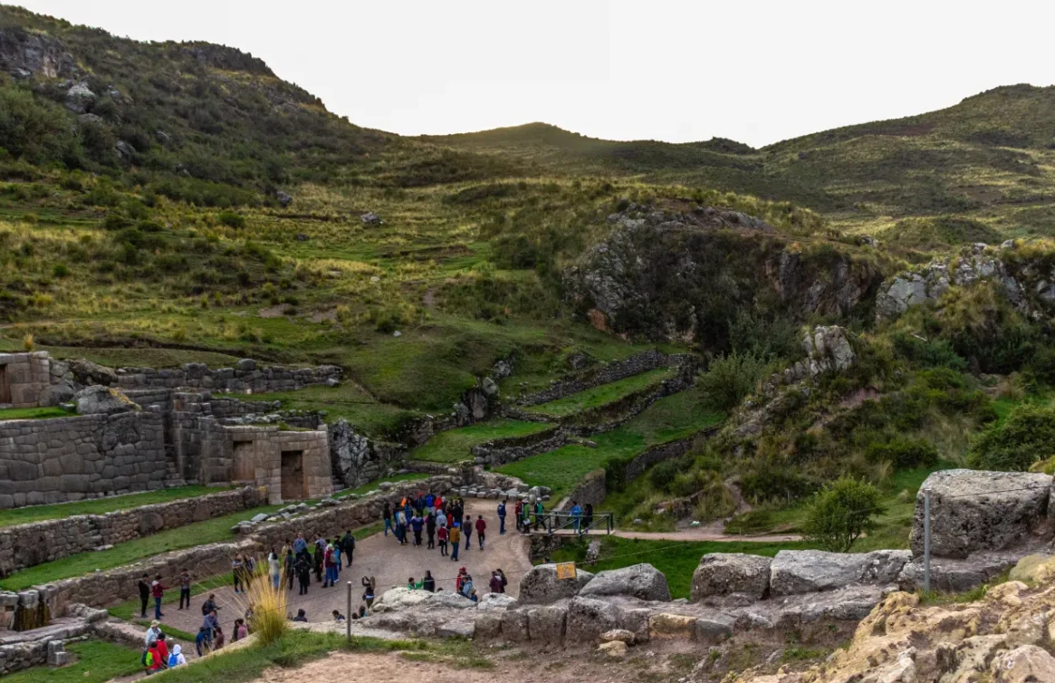 Um grupo de turistas visita as ruínas de Tambomachay entre as colinas verdes.