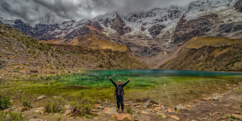 Turista comemorando em frente à Laguna Humantay, cercada por montanhas cobertas de neve.