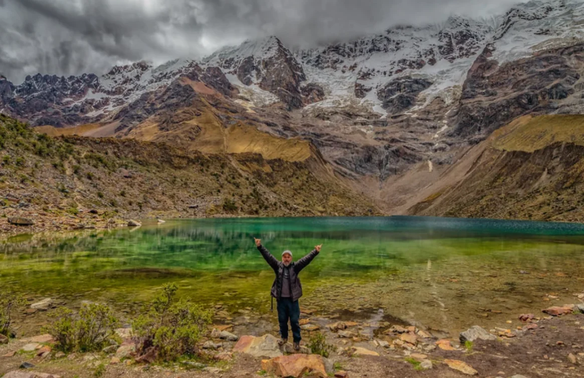 Turista comemorando em frente à Laguna Humantay, cercada por montanhas cobertas de neve.