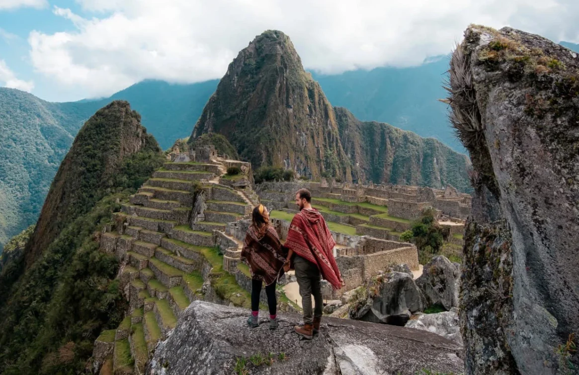 Dos Viajeros miran hacia el antiguo sitio arqueológico de Machu Picchu, rodeado de montañas y naturaleza.