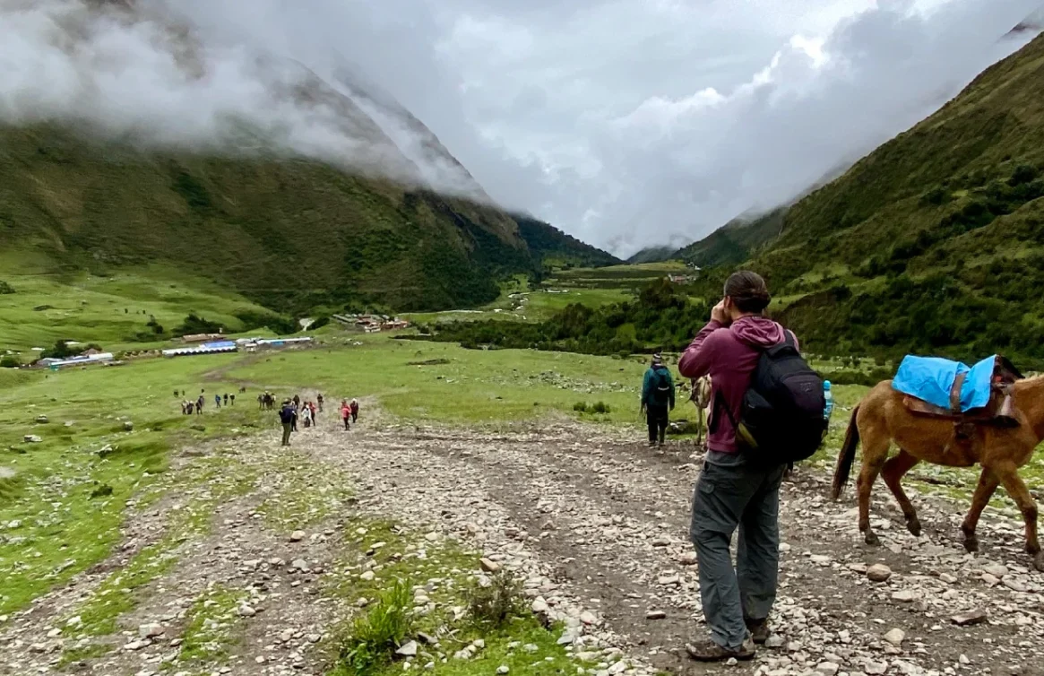 Turistas caminhando em direção ao Lago Humantay, com um cavalo e montanhas cobertas de neve ao fundo